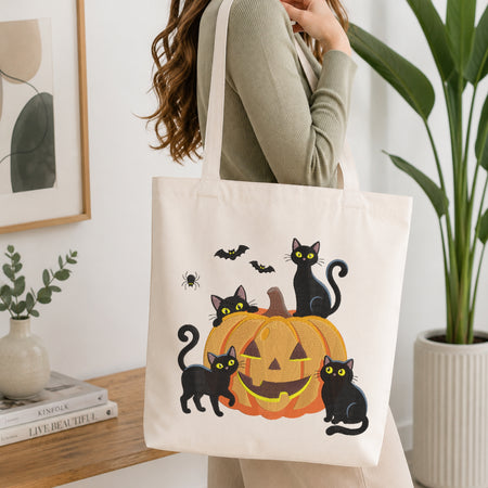 A person wearing a green long-sleeve top and carrying a natural canvas tote bag printed with the Halloween black cats and pumpkin machine embroidery design.