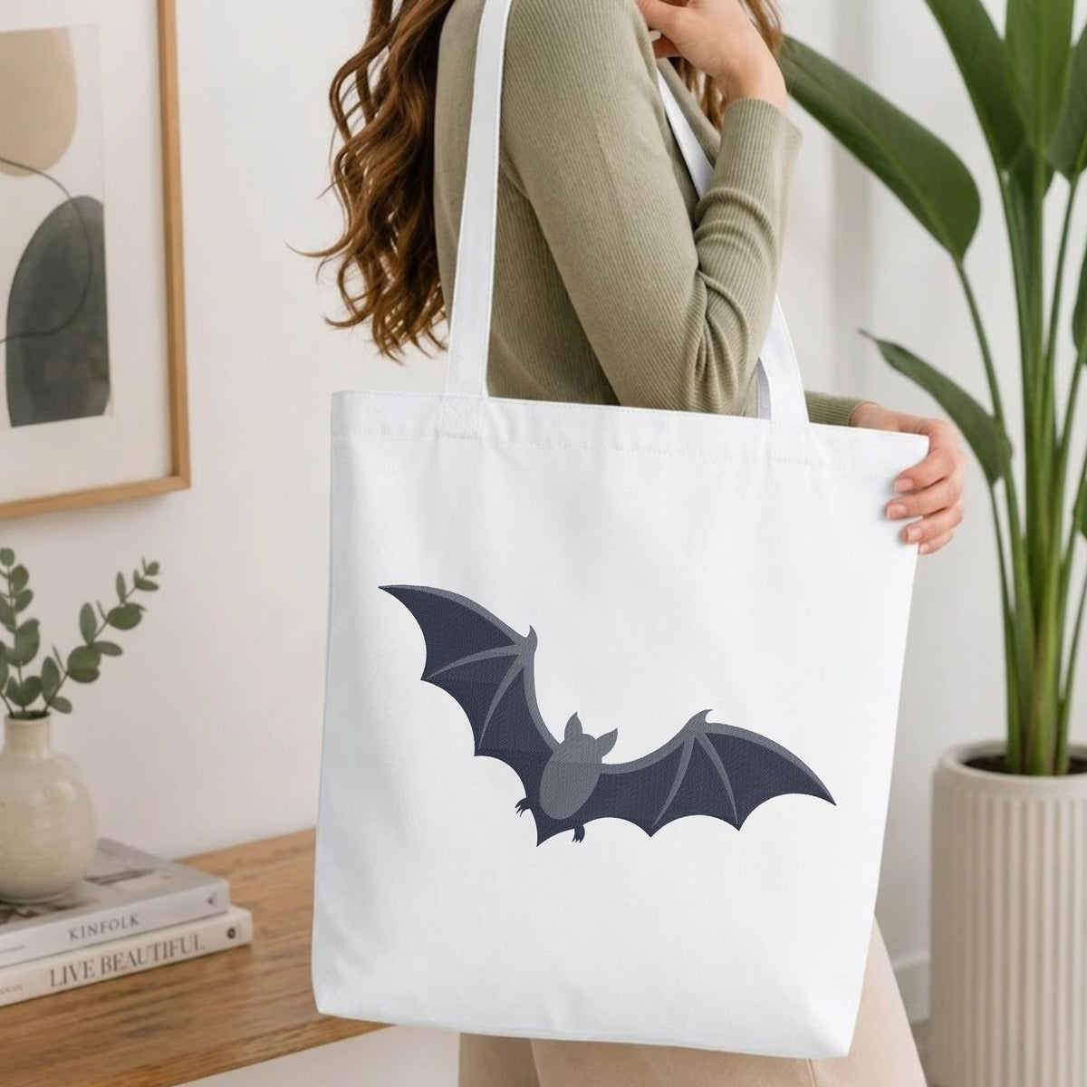 A woman wearing a sage green ribbed sweater carries a white canvas tote bag with a dark gray bat graphic printed on the front, set in a bright, modern room with a wooden table and potted plants.