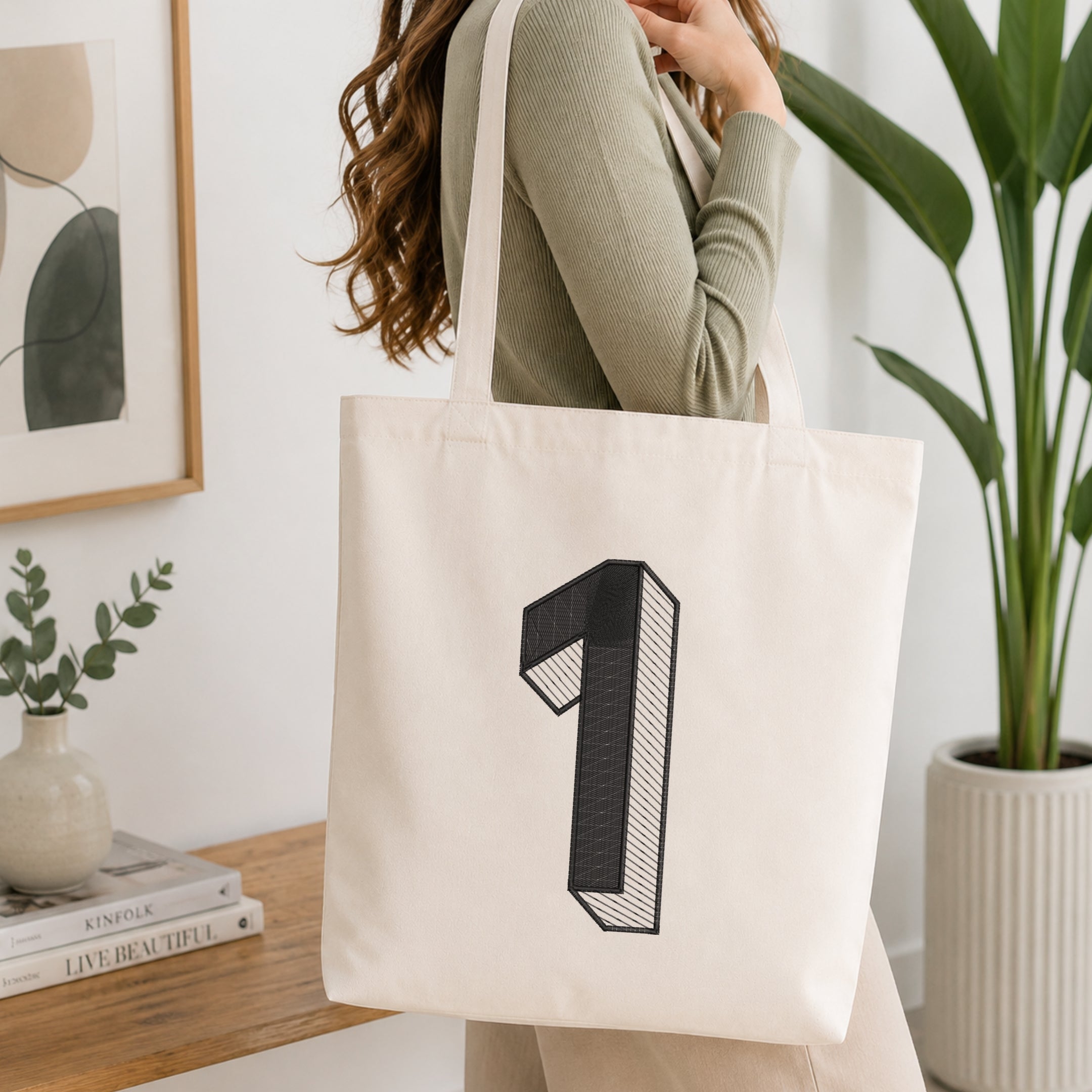 A woman wearing a sage green ribbed long-sleeve top carries a large off-white canvas tote bag with a black graphic of the number 1. The setting is a bright, modern room with a wooden table, books, a framed print, and a potted green plant.