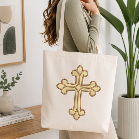 A woman wearing a sage green ribbed long-sleeve top carries a cream canvas tote bag with a large embroidered gold and brown cross on the front, set in a bright, modern room with a wooden table, books, and a potted plant.