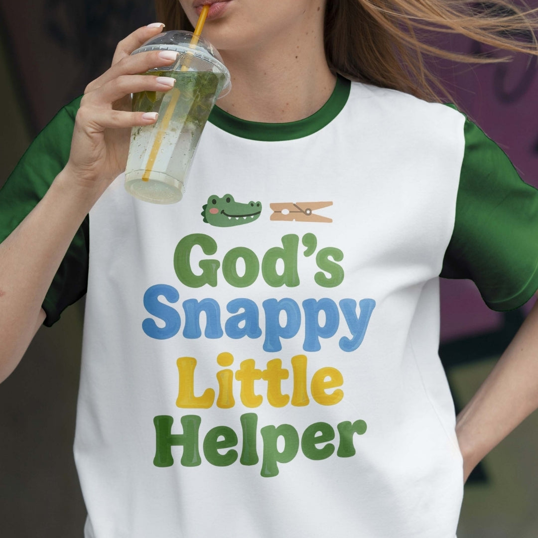 Person wearing a t-shirt with 'God's Snappy Little Helper' text and graphics, holding a drink.
