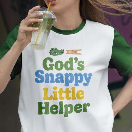 Person wearing a t-shirt with 'God's Snappy Little Helper' text and graphics, holding a drink.