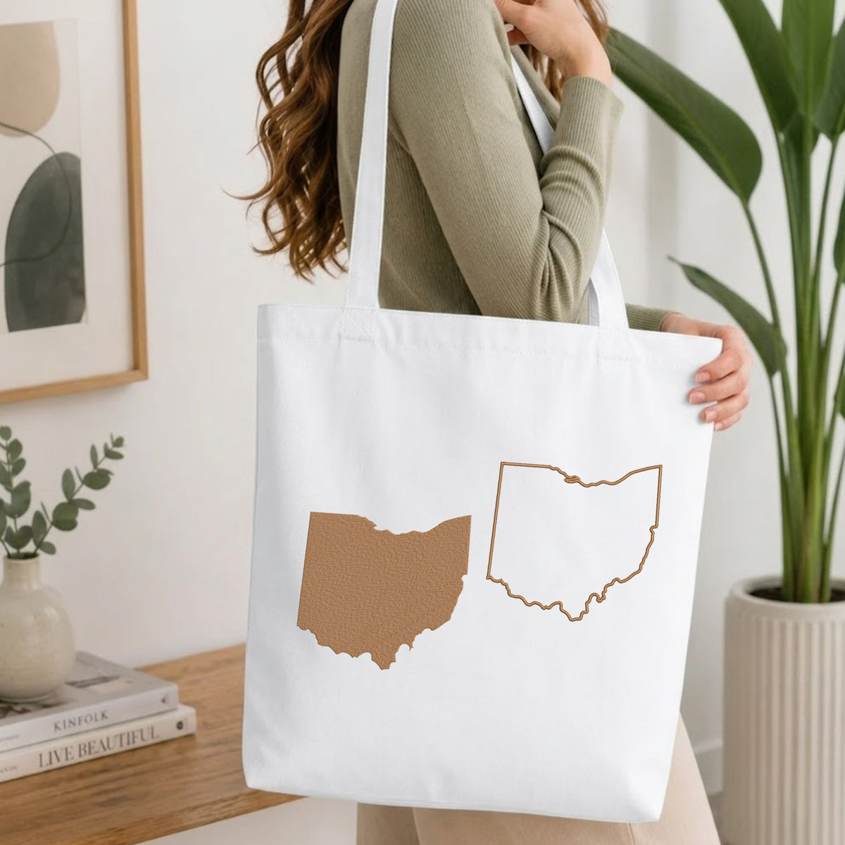 Woman carrying a white natural canvas tote bag featuring an embroidered Ohio state map design in a bright, modern interior setting.