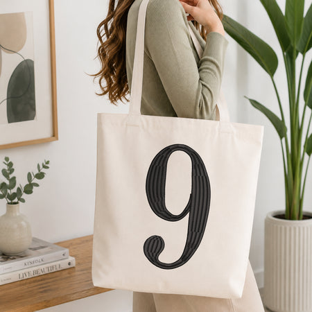 A woman wearing a sage green ribbed top carries a cream canvas tote bag with a large black textured number 9 printed on the front, set in a bright, modern room with a wooden console table and a potted plant.