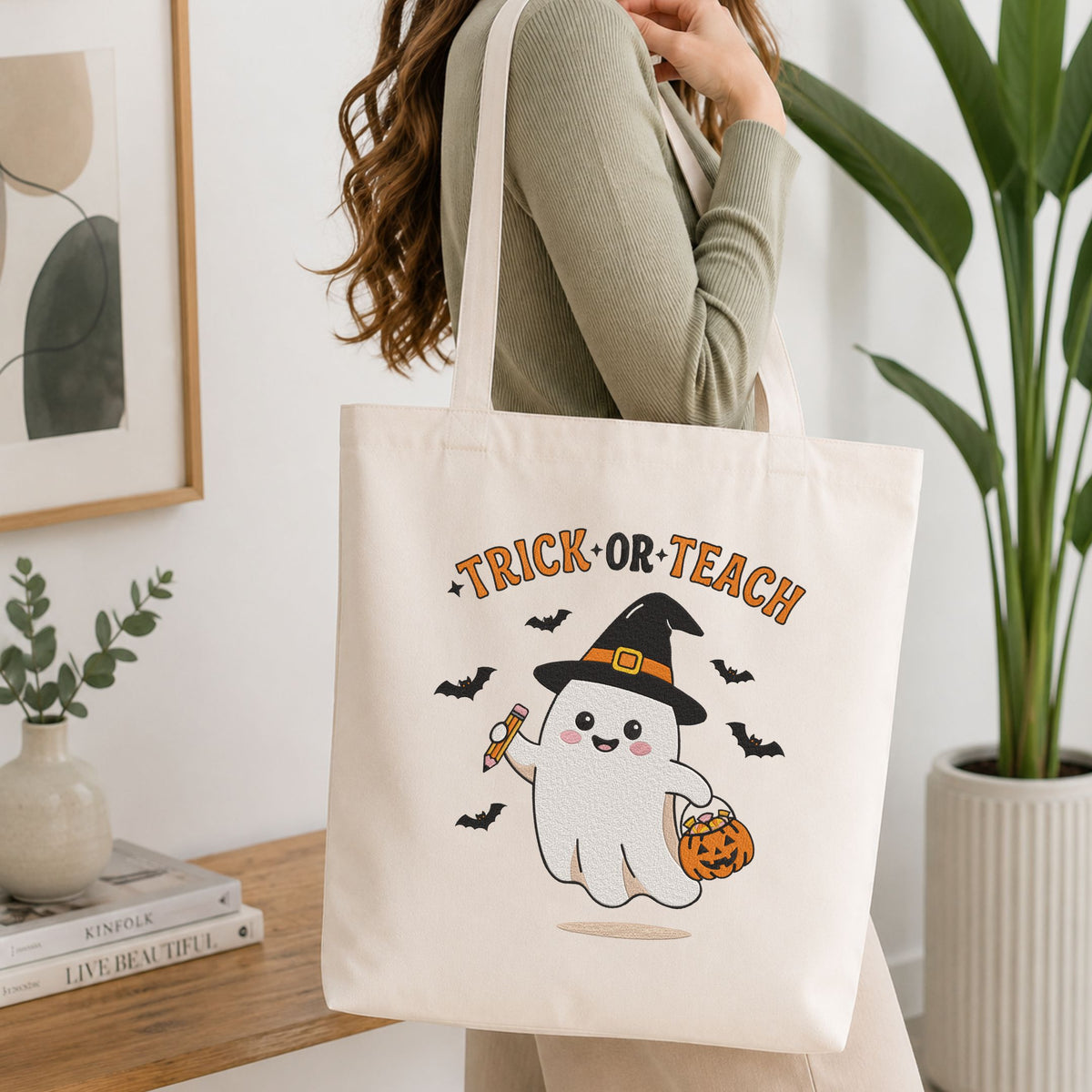 Woman carrying a white natural canvas tote bag featuring a "Trick or Teach" teacher ghost machine embroidery design in a bright, modern interior setting