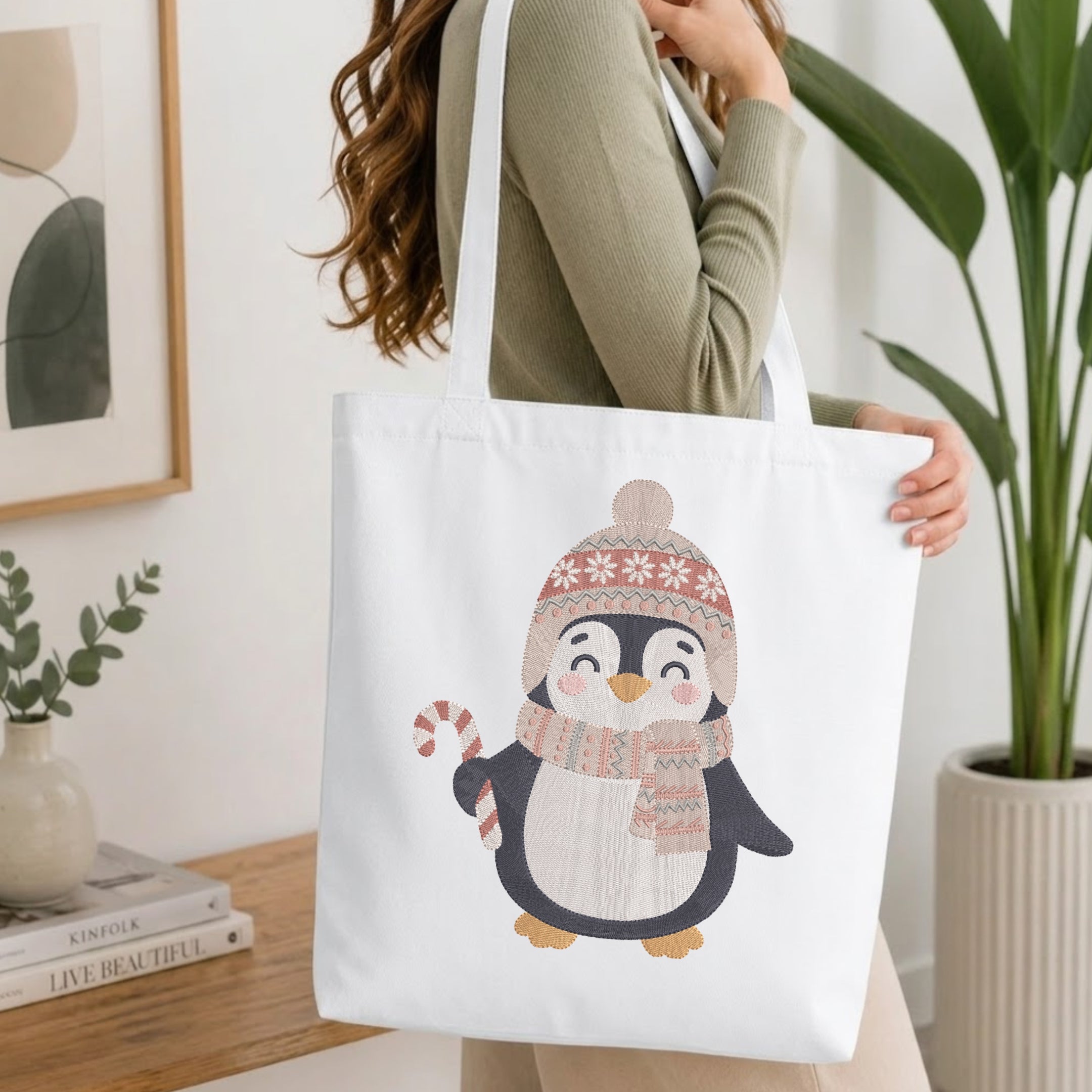 A woman wearing a sage green ribbed sweater carries a white canvas tote bag featuring a cute cartoon penguin wearing a pink and beige knit hat and scarf.