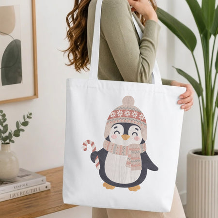 A woman wearing a sage green ribbed sweater carries a white canvas tote bag featuring a cute cartoon penguin wearing a pink and beige knit hat and scarf.