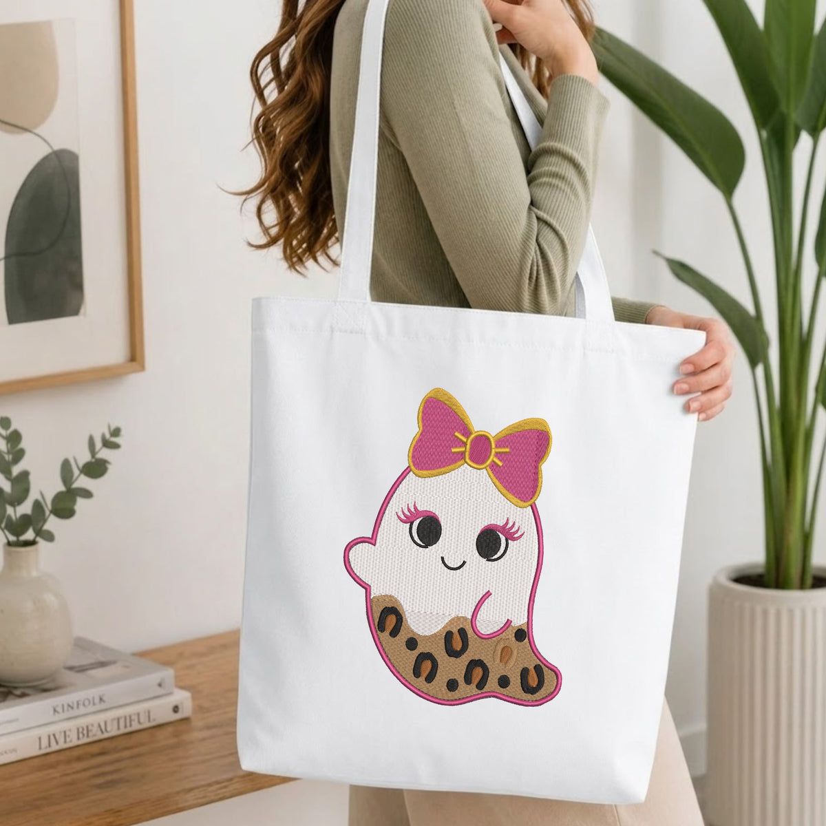 Woman carrying a white natural canvas tote bag featuring a cute leopard print ghost embroidery design in a bright, modern interior setting.