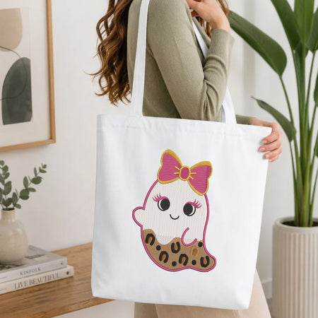 Woman carrying a white natural canvas tote bag featuring a cute leopard print ghost embroidery design in a bright, modern interior setting.