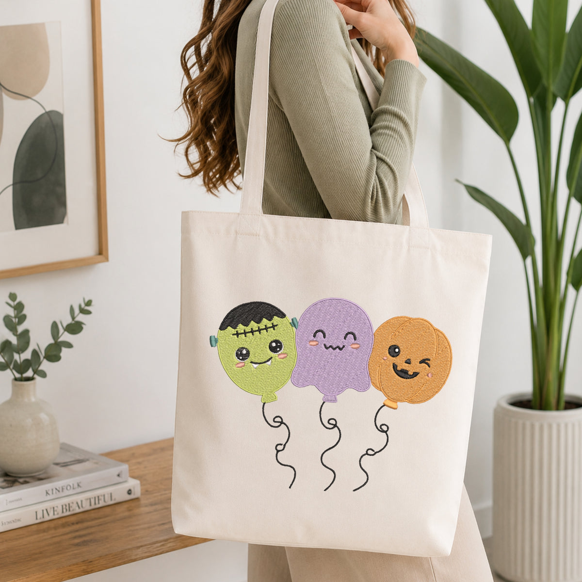 An off-white canvas tote bag with three cute embroidered Halloween balloon characters (Frankenstein's monster, purple ghost, and orange pumpkin) being carried on a woman's shoulder in a bright, modern room.