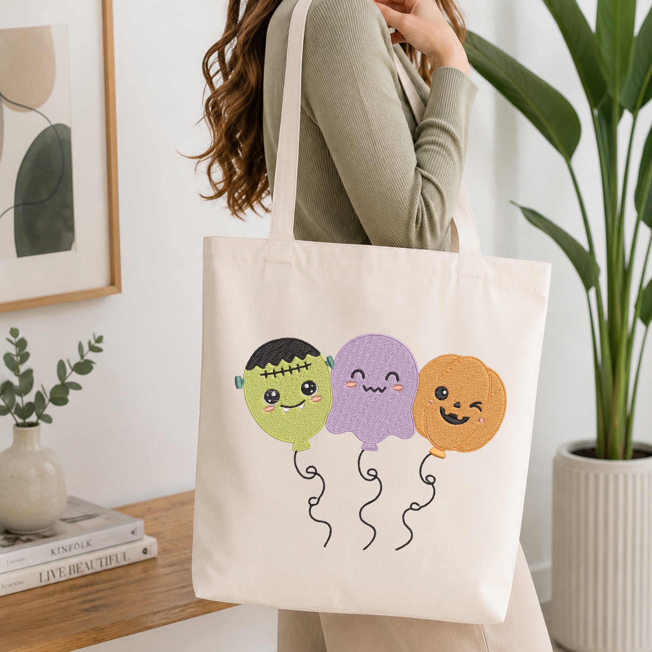 An off-white canvas tote bag with three cute embroidered Halloween balloon characters (Frankenstein's monster, purple ghost, and orange pumpkin) being carried on a woman's shoulder in a bright, modern room.