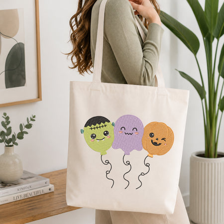 An off-white canvas tote bag with three cute embroidered Halloween balloon characters (Frankenstein's monster, purple ghost, and orange pumpkin) being carried on a woman's shoulder in a bright, modern room.