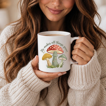 A woman in a cream knit sweater holds a white ceramic mug with a colorful mushroom illustration and the phrase 'He makes all things grow' in a cozy indoor setting.