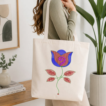 A woman wearing a sage green ribbed long-sleeve top carries a cream canvas tote bag with a colorful embroidered floral design. The background shows a cozy indoor setting with a wooden table, books, a small vase, and a potted plant.