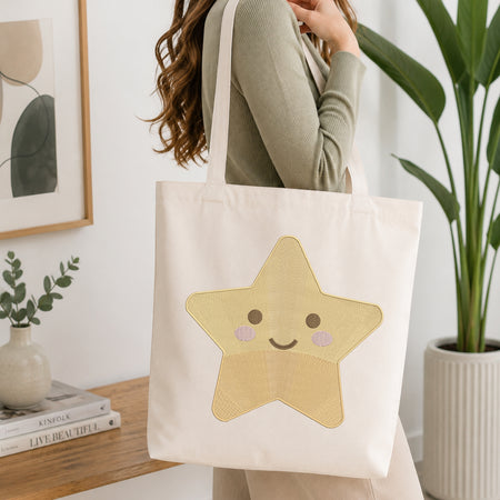 A person wearing a sage green ribbed top carries a cream canvas tote bag with a large embroidered yellow star face on the front, set in a bright, modern room with a wooden table, framed art, and a potted plant.