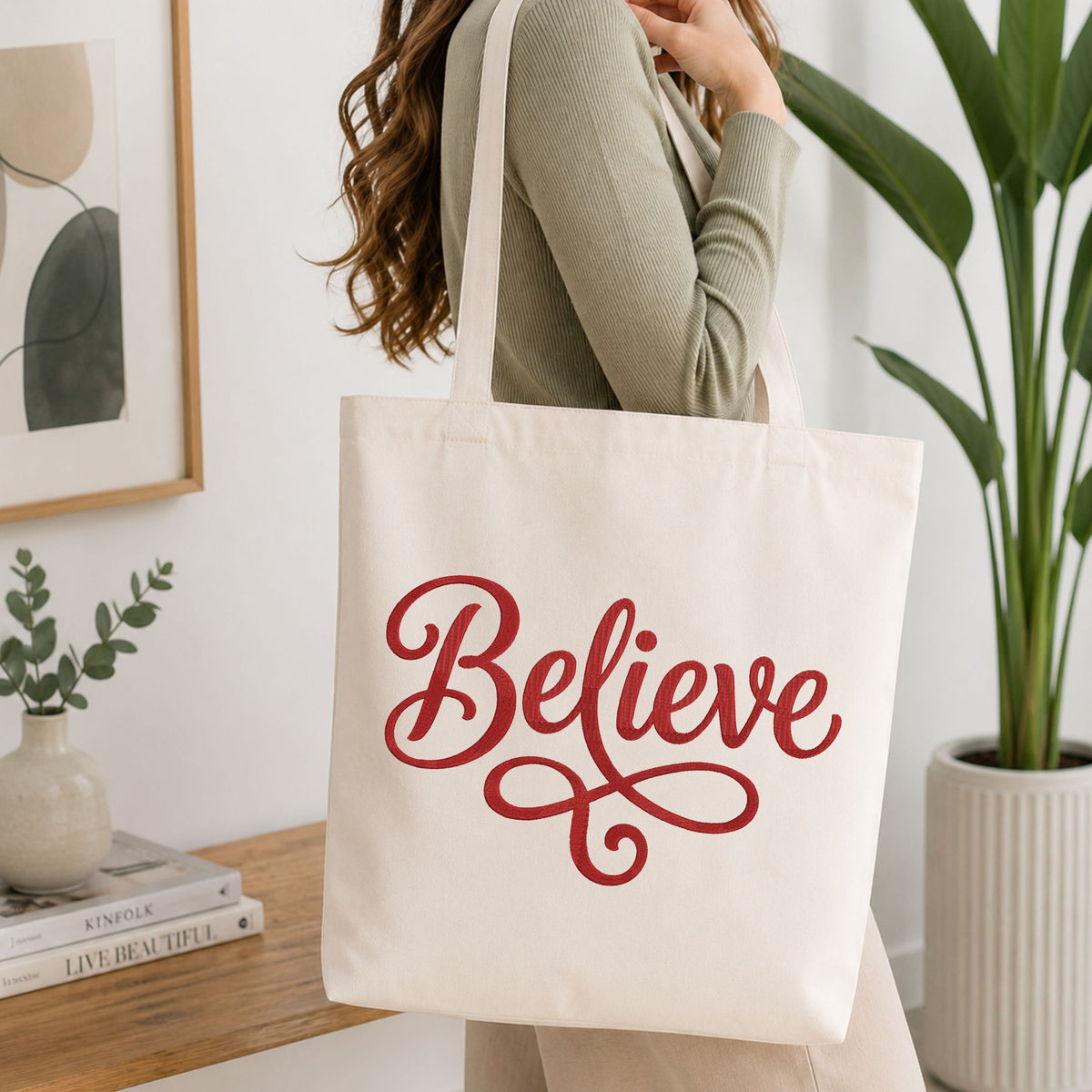 Woman carrying a natural canvas tote bag featuring an embroidered "Believe" lettering design in a bright, modern interior setting.
