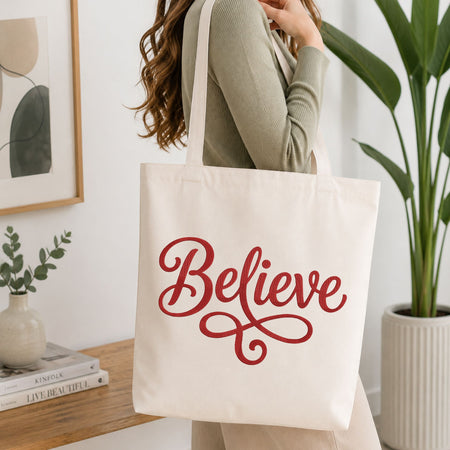 Woman carrying a natural canvas tote bag featuring an embroidered "Believe" lettering design in a bright, modern interior setting.