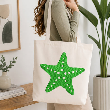 An off-white canvas tote bag with a large green starfish applique on the front, carried by a woman in a ribbed sage green top in a bright, modern room.