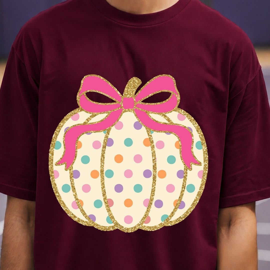 A person wearing a burgundy short-sleeve T-shirt with a large pumpkin graphic on the chest. The pumpkin is cream-colored with multicolored polka dots and a pink bow with gold glitter outlines.