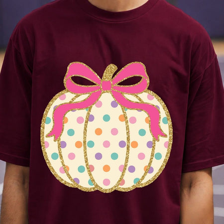 A person wearing a burgundy short-sleeve T-shirt with a large pumpkin graphic on the chest. The pumpkin is cream-colored with multicolored polka dots and a pink bow with gold glitter outlines.