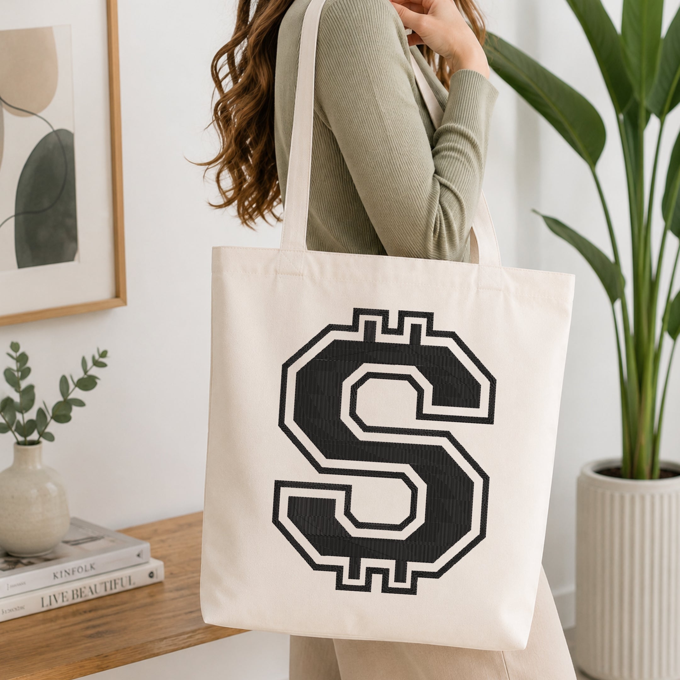 A woman wearing a sage green ribbed long-sleeve top carries a cream canvas tote bag with a large black and white dollar sign graphic on the front, set in a bright, modern room with a wooden console table and a potted plant.
