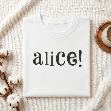 A folded white t-shirt with the word 'alice!' printed in black serif font, styled on a textured cream knit blanket with cotton bolls and a cup in the background.
