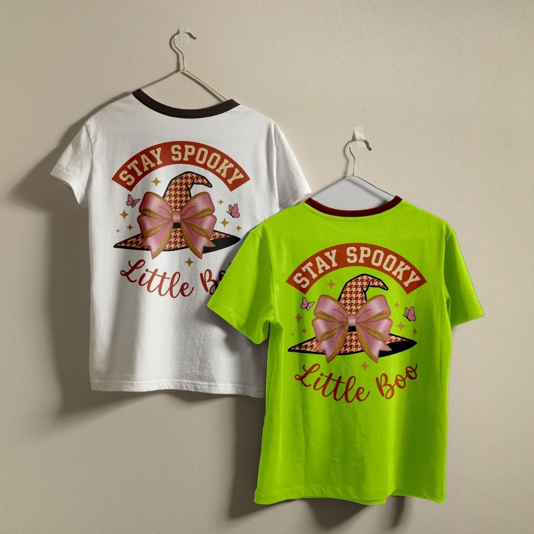 Two children's short-sleeve ringer T-shirts hanging on hangers against a plain beige wall. The left shirt is white with a dark red collar and shows the back graphic: a witch hat with a pink bow, small stars, and the words 'STAY SPOOKY Little Boo.