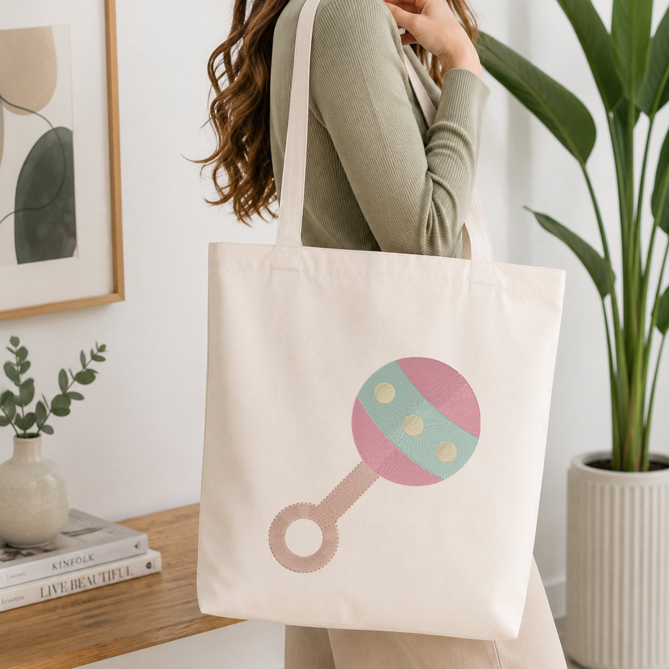 A woman wearing a sage green ribbed long-sleeve top carries a cream canvas tote bag with a pink and mint green rattle graphic. The scene is set in a bright, modern room with a wooden shelf, framed art, a small potted plant, and a larger green plant in the background.