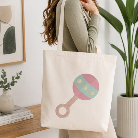A woman wearing a sage green ribbed long-sleeve top carries a cream canvas tote bag with a pink and mint green rattle graphic. The scene is set in a bright, modern room with a wooden shelf, framed art, a small potted plant, and a larger green plant in the background.