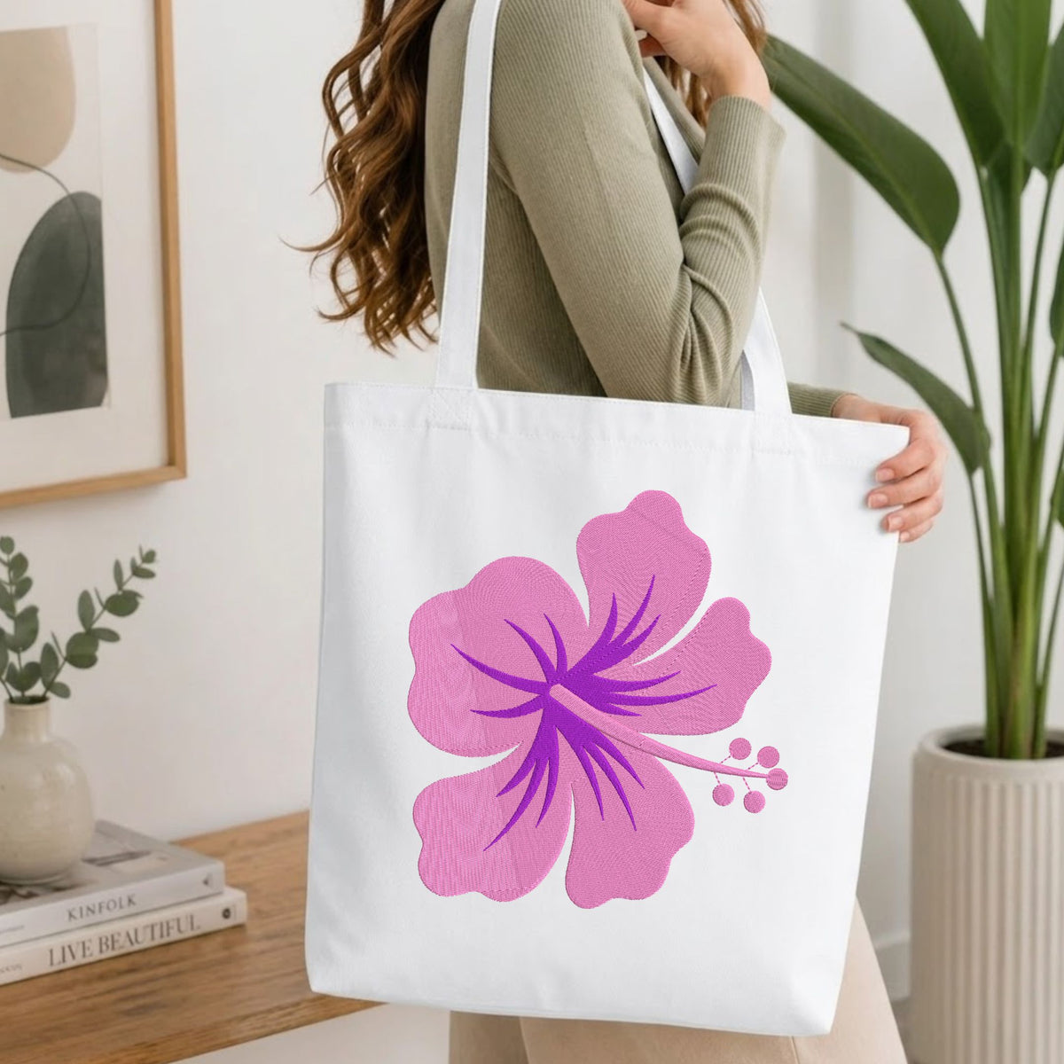 A woman wearing a sage green ribbed long-sleeve top carries a white canvas tote bag with a large pink hibiscus flower graphic on the front, set in a bright, modern room with a wooden table and potted plants.