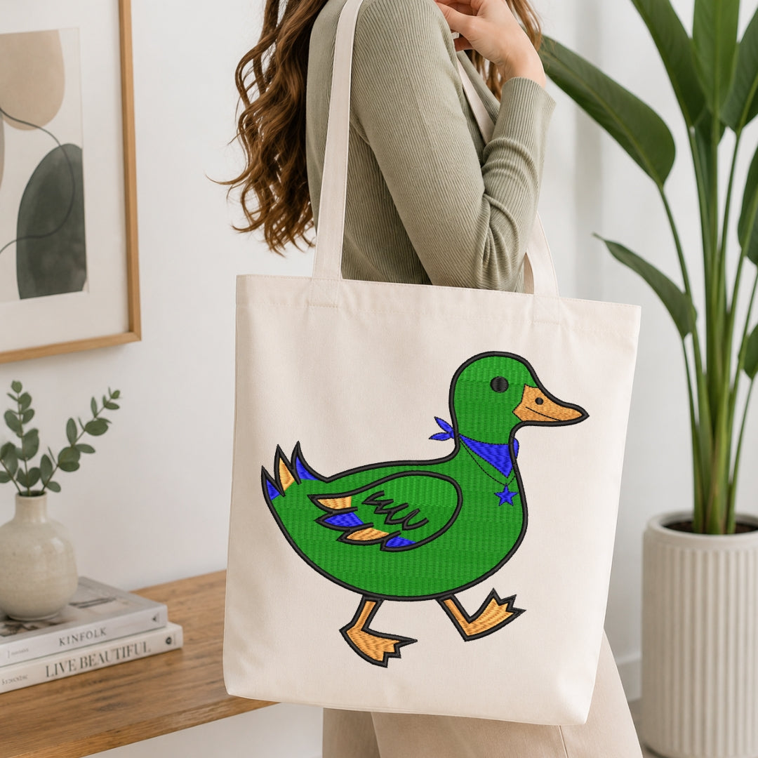 A woman wearing a sage green ribbed sweater carries a cream canvas tote bag featuring a colorful embroidered-style green duck with yellow-orange beak and feet, and a blue bow tie, in a bright, modern room with a potted plant and books.
