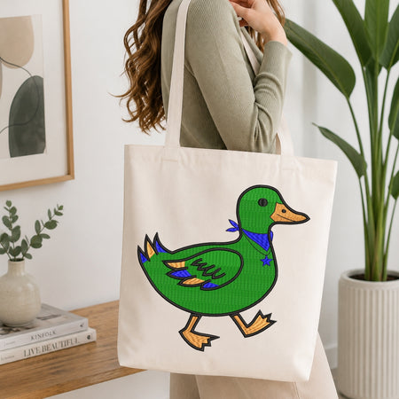 A woman wearing a sage green ribbed sweater carries a cream canvas tote bag featuring a colorful embroidered-style green duck with yellow-orange beak and feet, and a blue bow tie, in a bright, modern room with a potted plant and books.
