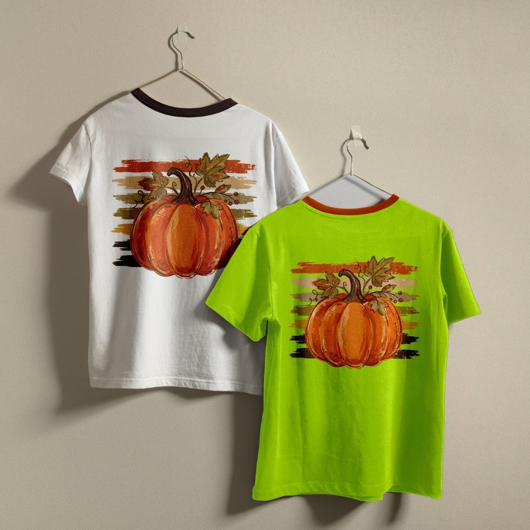 Two children's short-sleeve t-shirts hanging on hangers against a plain beige wall. The left shirt is white with a dark brown collar and features a large orange pumpkin graphic on the back with a sunset-style stripe background.