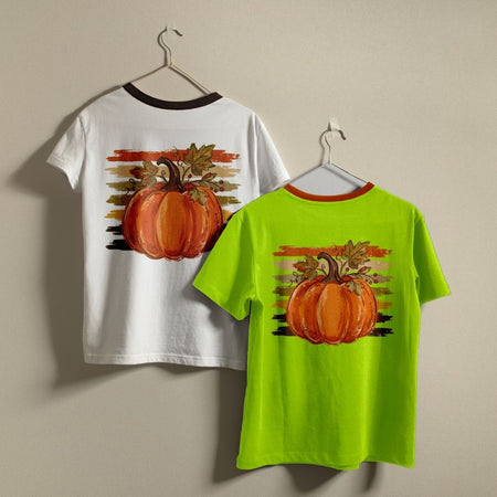 Two children's short-sleeve t-shirts hanging on hangers against a plain beige wall. The left shirt is white with a dark brown collar and features a large orange pumpkin graphic on the back with a sunset-style stripe background.