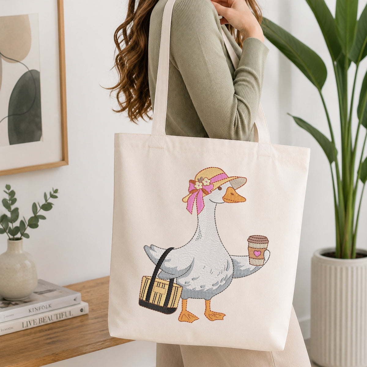 A woman in a sage green ribbed sweater carries a cream canvas tote bag with a cartoon goose holding a coffee cup and a striped bag. The scene is set in a bright, modern room with a wooden table, books, a framed print, and a potted plant.