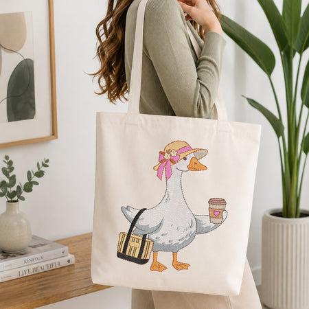 A woman in a sage green ribbed sweater carries a cream canvas tote bag with a cartoon goose holding a coffee cup and a striped bag. The scene is set in a bright, modern room with a wooden table, books, a framed print, and a potted plant.