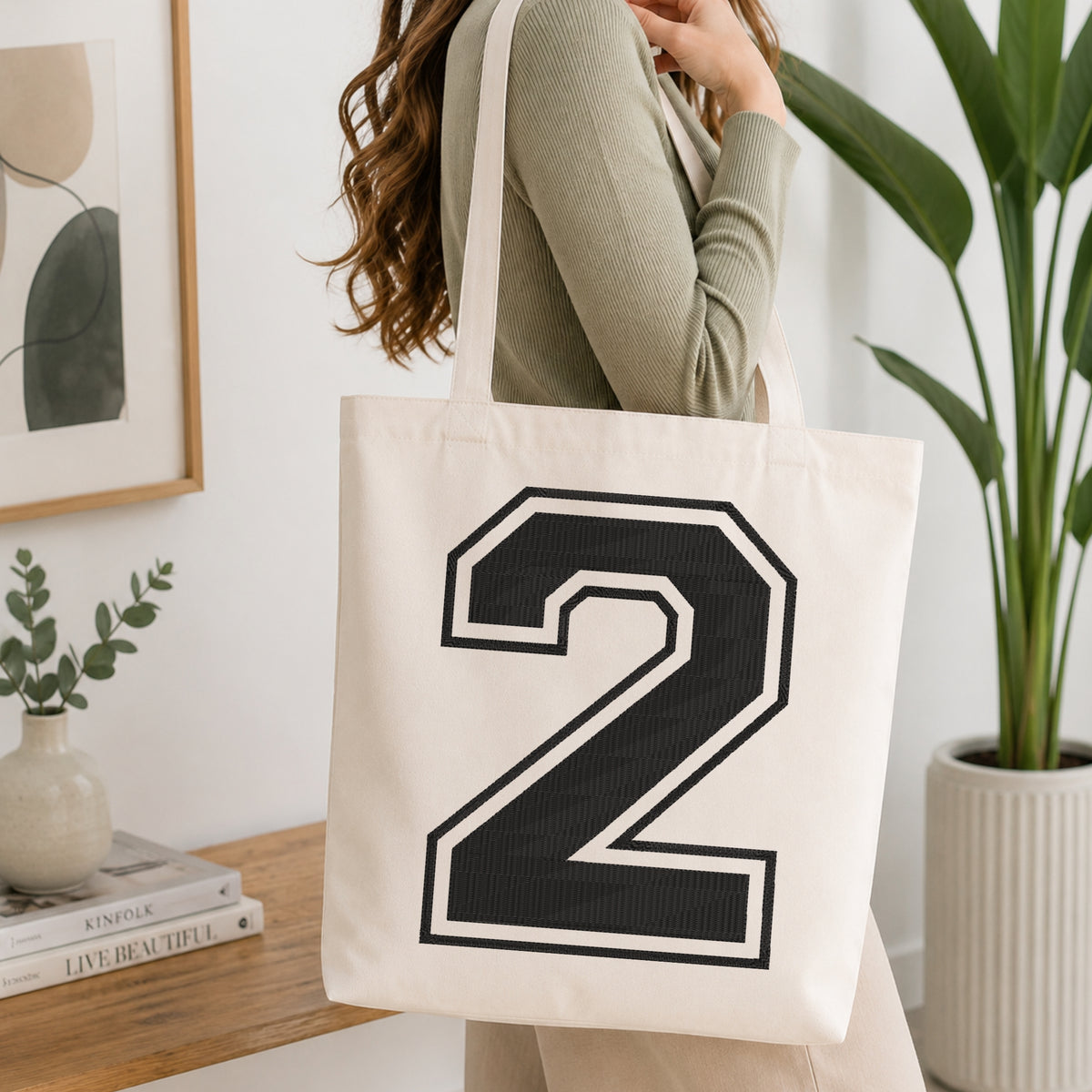 A woman wearing a sage green ribbed long-sleeve top carries a cream canvas tote bag with a large black and white number 2 printed on the front, set in a bright, modern room with a wooden console table and a potted plant.