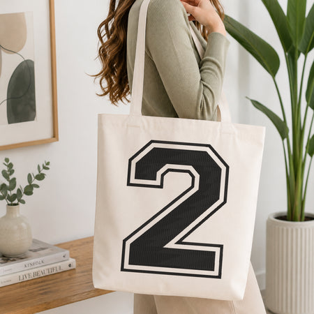 A woman wearing a sage green ribbed long-sleeve top carries a cream canvas tote bag with a large black and white number 2 printed on the front, set in a bright, modern room with a wooden console table and a potted plant.