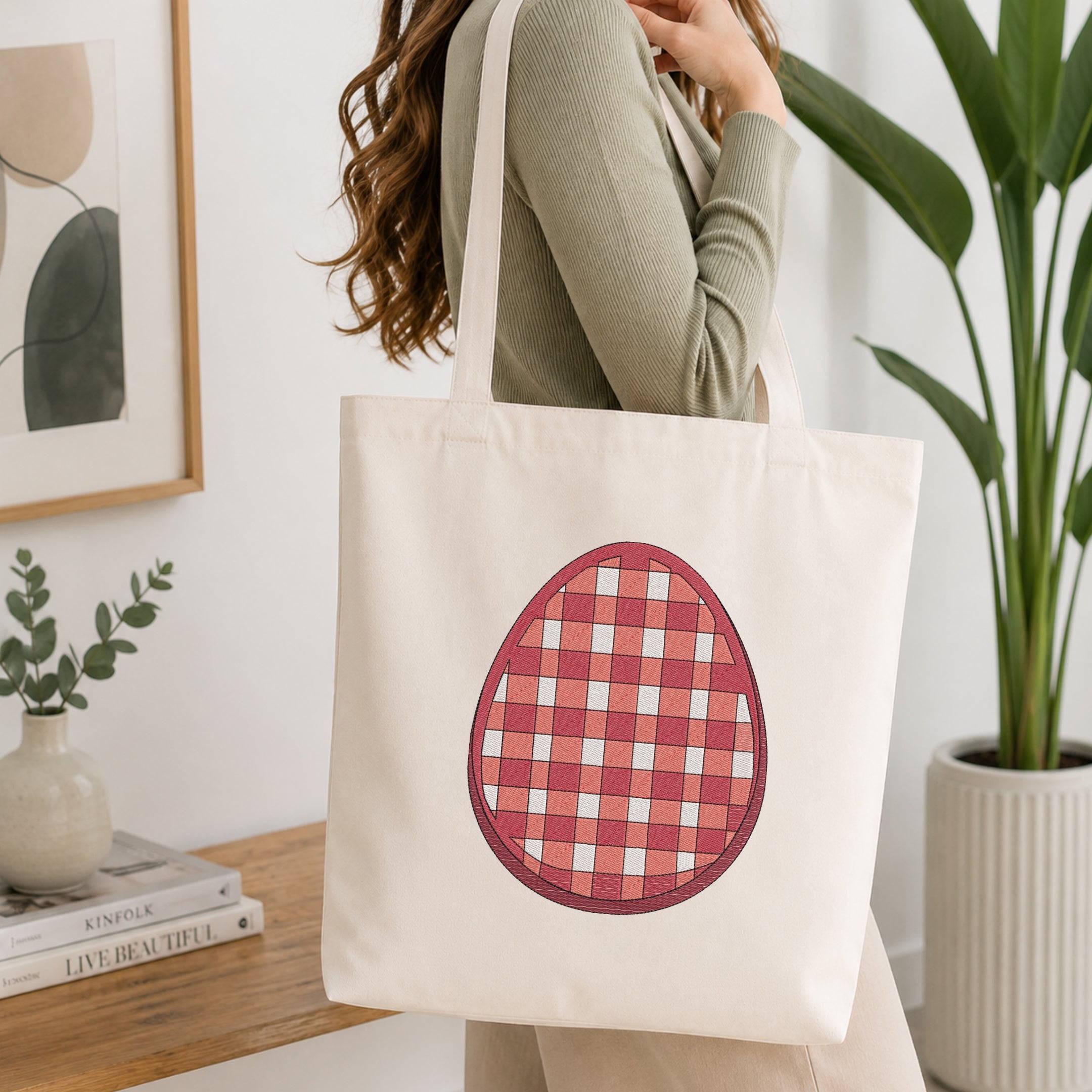 A woman wearing a sage green ribbed long-sleeve top carries a cream canvas tote bag with a red and white gingham checkered egg graphic on the front, set in a bright, modern room with a wooden table, framed art, and a potted plant.