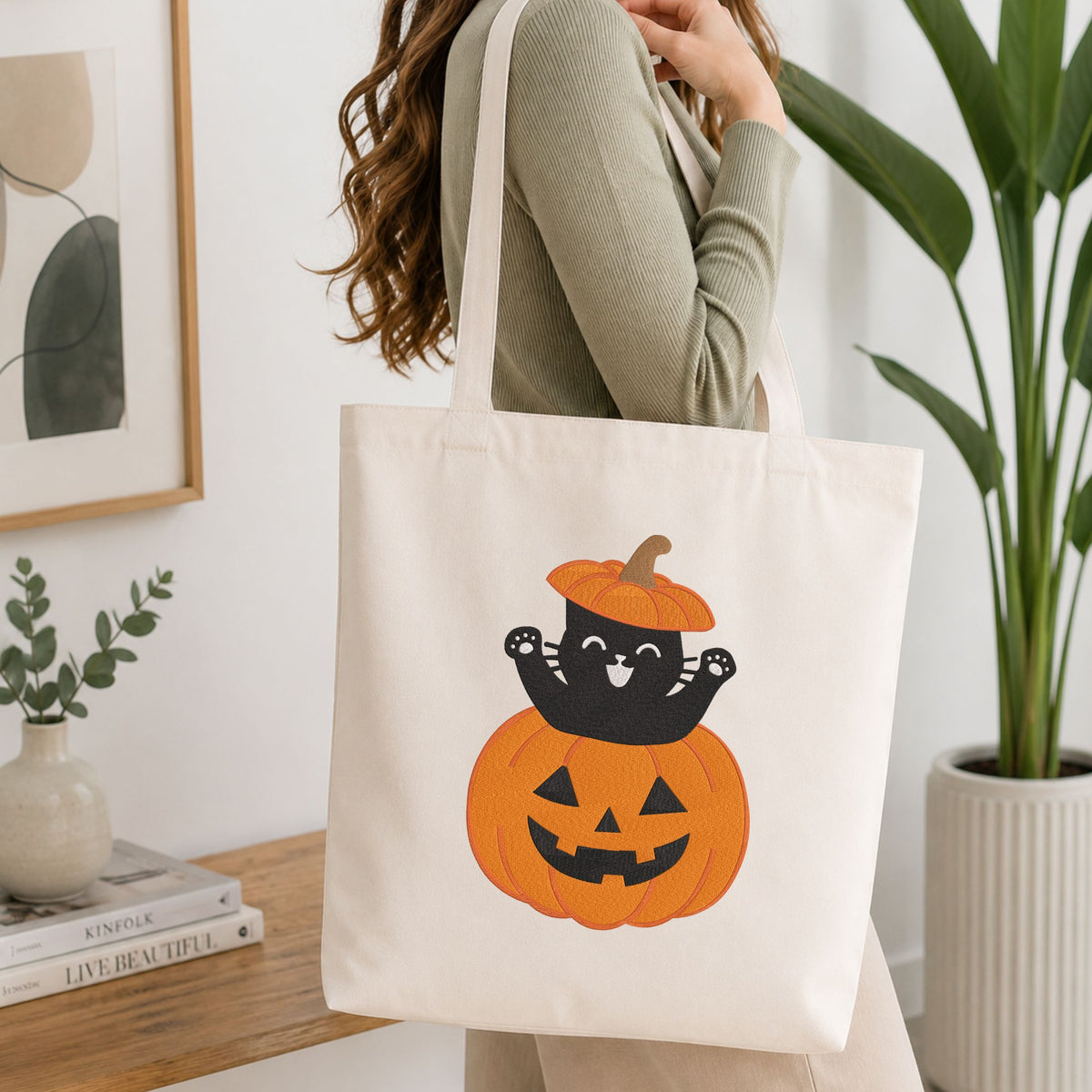 Woman carrying a white natural canvas tote bag featuring a cute black cat wearing a pumpkin hat sitting inside a jack-o-lantern machine embroidery design in a bright, modern interior setting