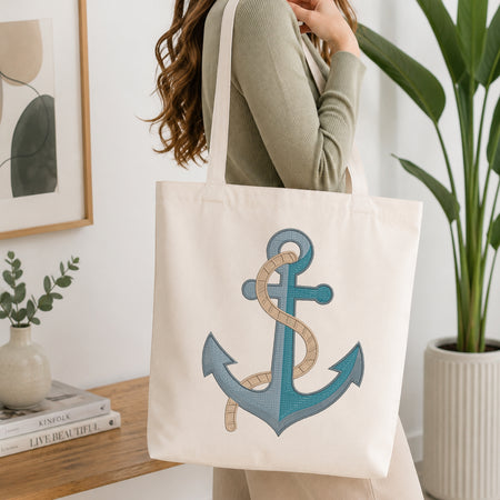 A woman wearing a sage green ribbed sweater carries a cream canvas tote bag with a large embroidered anchor design in shades of blue and beige. The scene is set in a bright, modern room with a wooden table, framed art, a small plant, and a potted green plant in the background.