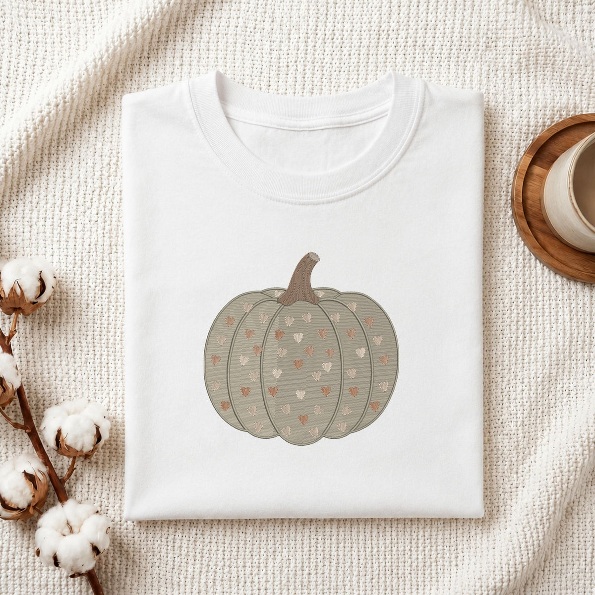 A folded white crewneck t-shirt featuring a graphic of a striped gray-green pumpkin with small pink and beige hearts, styled on a textured cream knit blanket with cotton bolls and a wooden cup in the background.