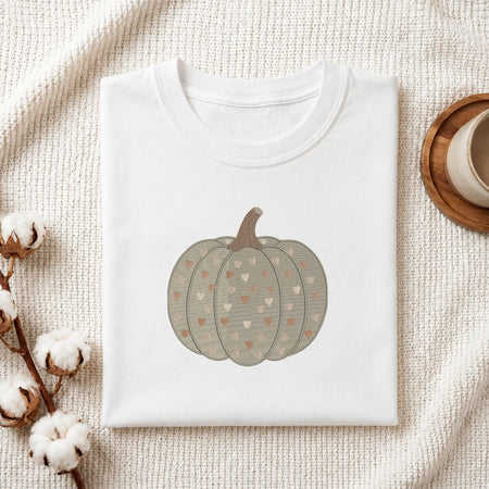 A folded white crewneck t-shirt featuring a graphic of a striped gray-green pumpkin with small pink and beige hearts, styled on a textured cream knit blanket with cotton bolls and a wooden cup in the background.