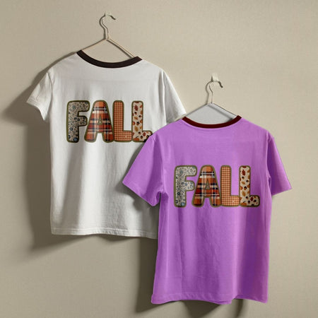 Two short-sleeve T-shirts hanging on hangers against a plain beige wall. The back of one white shirt and the front of one purple shirt both display a large patchwork-style 'FALL' graphic in earthy plaid and leopard print tones.