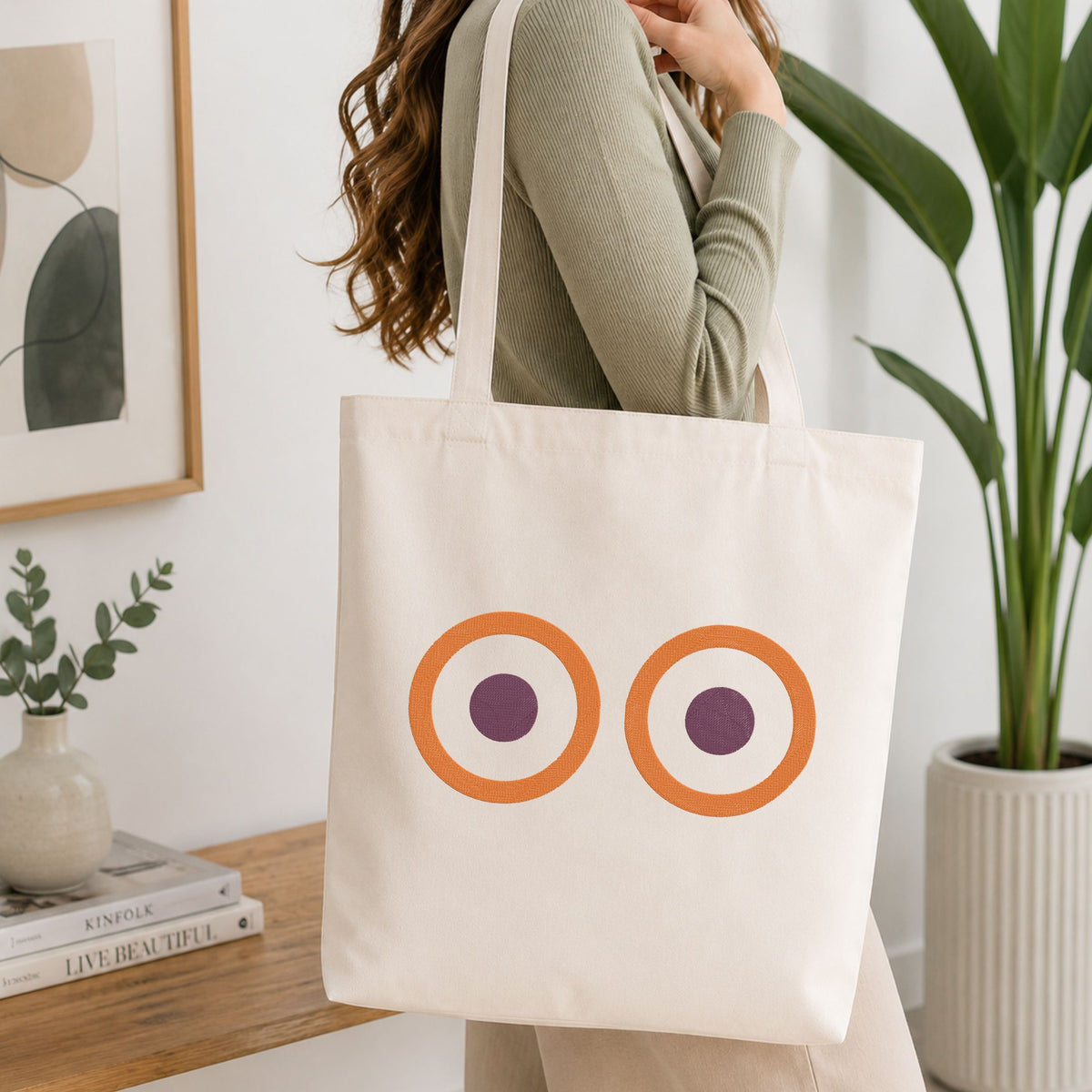 A woman wearing a sage green ribbed long-sleeve top carries a cream canvas tote bag featuring a graphic of two orange rings with purple centers, set against a bright, modern room with a wooden table, books, and a potted plant.