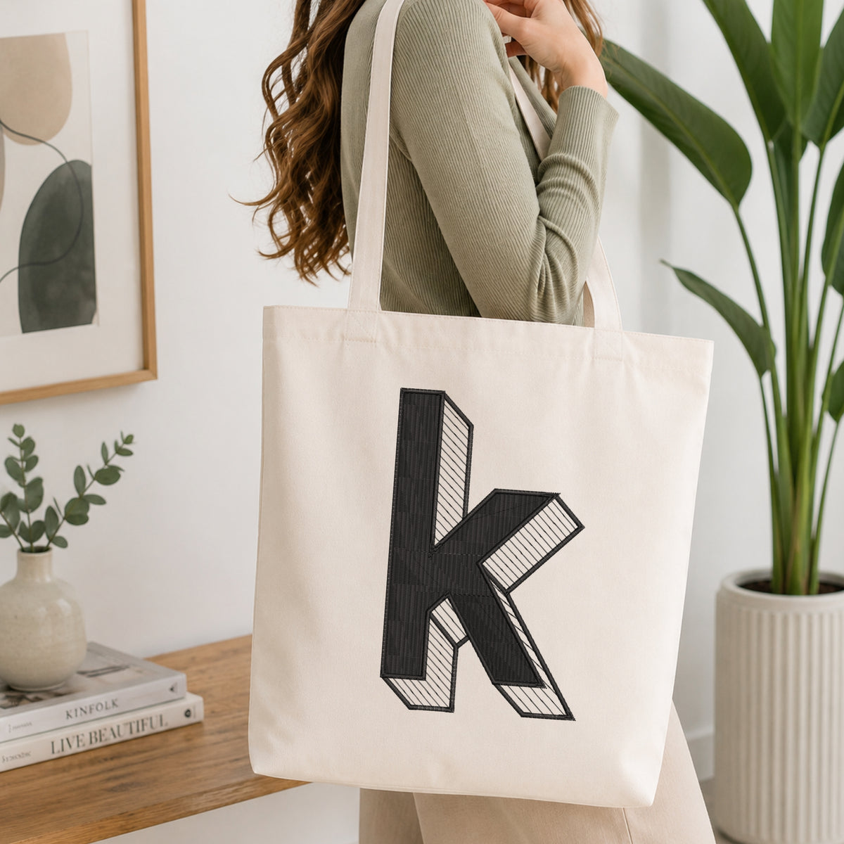 A woman wearing a sage green ribbed long-sleeve top carries a cream canvas tote bag with a large black 3D-effect letter 'K' printed on the front, set in a bright, modern room with a wooden table, framed art, and a potted plant.