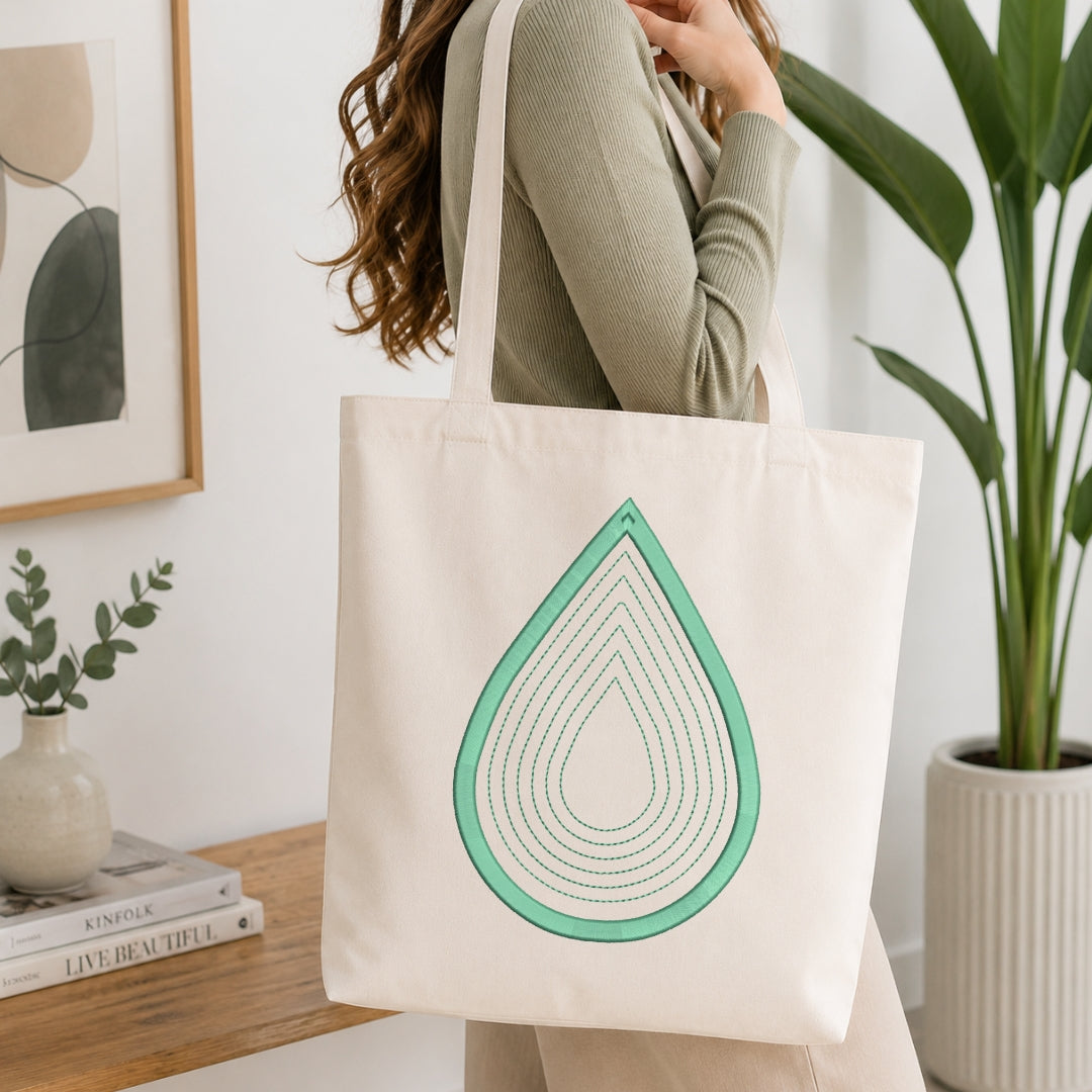 A woman wearing a sage green ribbed sweater carries a cream canvas tote bag with a mint green teardrop graphic design. The scene is set in a bright, modern room with a wooden table, books, a framed print, and a potted plant.
