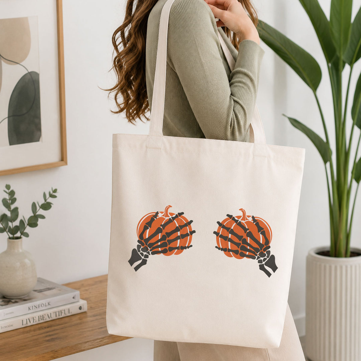 Woman carrying a white natural canvas tote bag featuring a skeleton hands holding pumpkins machine embroidery design in a bright, modern interior setting   