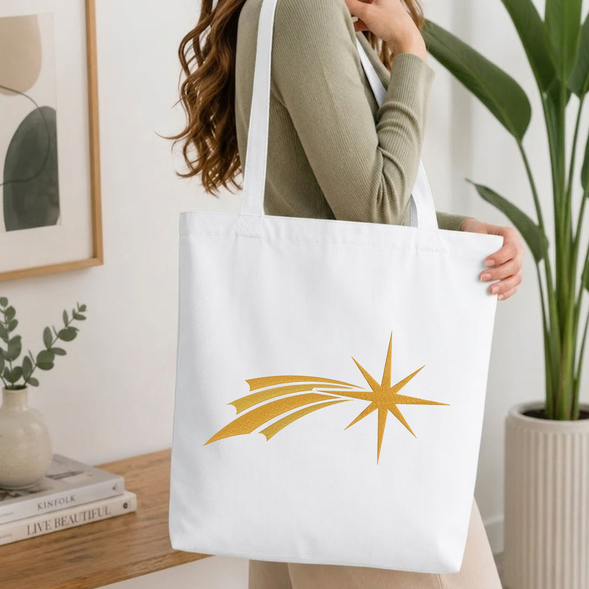 Woman carrying a white natural canvas tote bag featuring an embroidered golden comet star design in a bright, modern interior setting