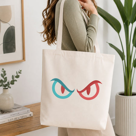 A woman wearing a sage green ribbed sweater carries a cream canvas tote bag with a graphic of two angry eyes, one in teal and one in red, in a bright, modern room.